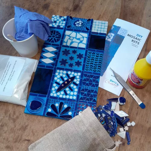 mosaic kit contents on a wooden table. finsihed msoaic wall art surrounded by ceramic and glass tiles, bag of grout powder, tools and instructions leaflet.
